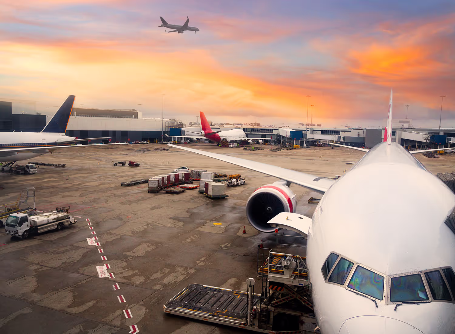 Airplane parked at airport gate, with another plane taking off against a colorful sunset sky.
