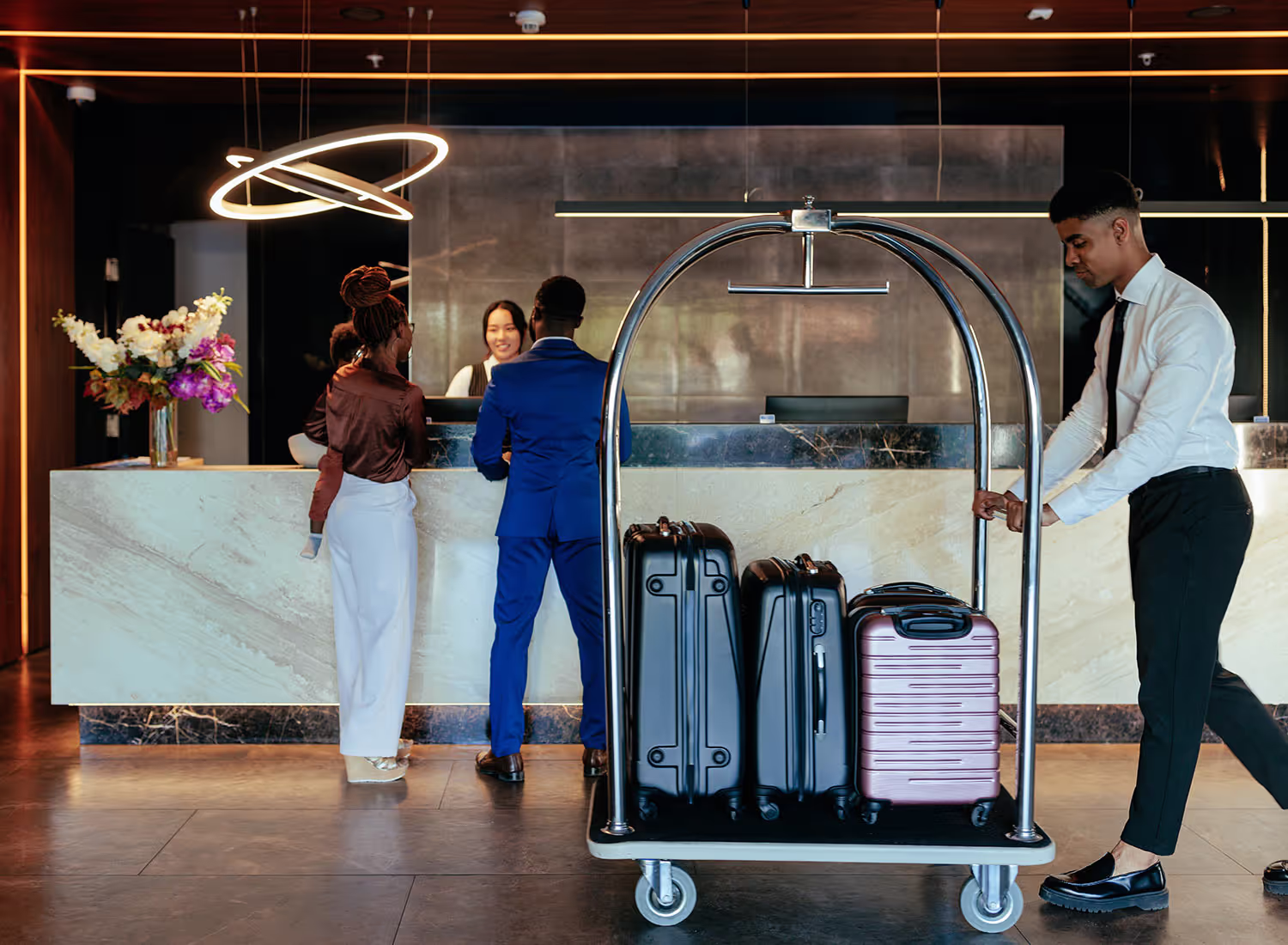 A hotel staff member pushes a luggage cart as a couple checks in at the front desk.
